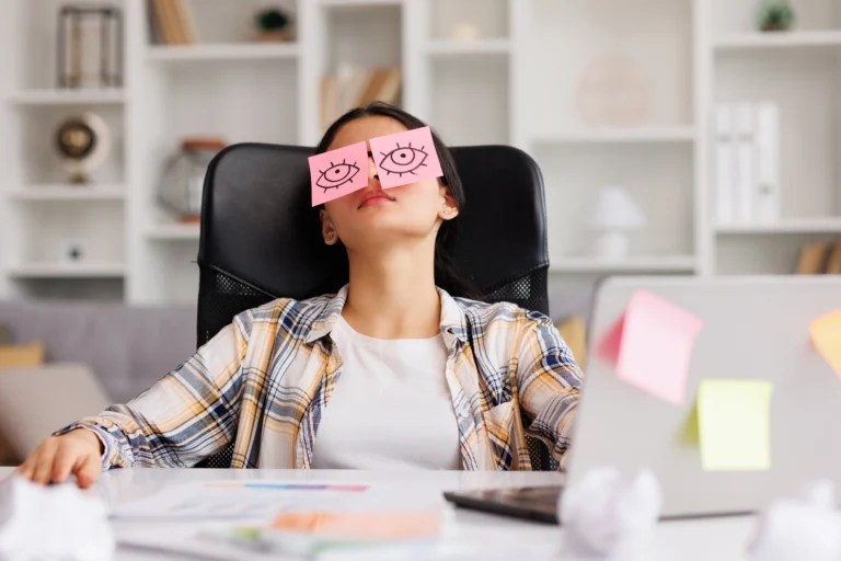 Kobieta, próbująca ukoić zaczerwienione oczy, siedzi na krześle za biurkiem w pracy z naklejonymi na oczy różowymi karteczkami post-it z narysowanymi markerem oczami.