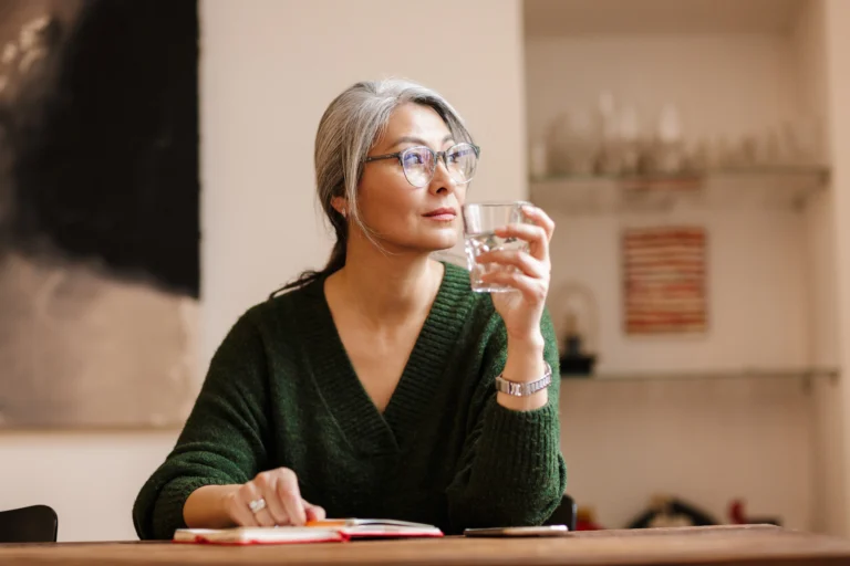 Kobieta w wieku dojrzałym pije wodę podczas notowania w kalendarzu, pamiętając o dobrych nawykach, poprawiających nawilżenie oczu.