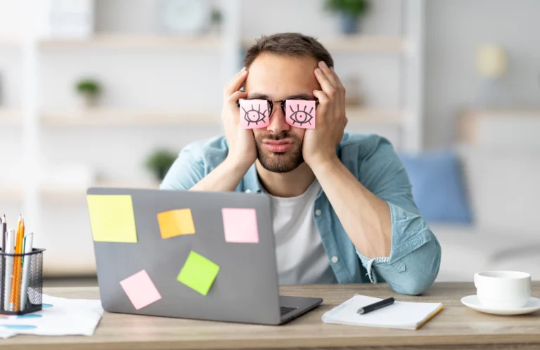 Mężczyzna siedzący przed laptopem szuka sposobów na zaczerwienione oczy, przyklejając sobie karteczki post-it do okularów.